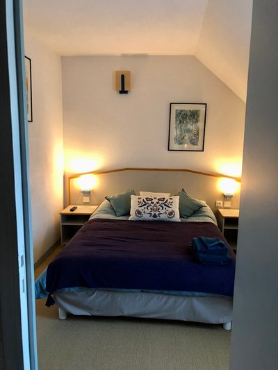 Chambre Campanule : lit double sous rampant, couvre-lit bleu nuit, deux lampes de chevet allumées.